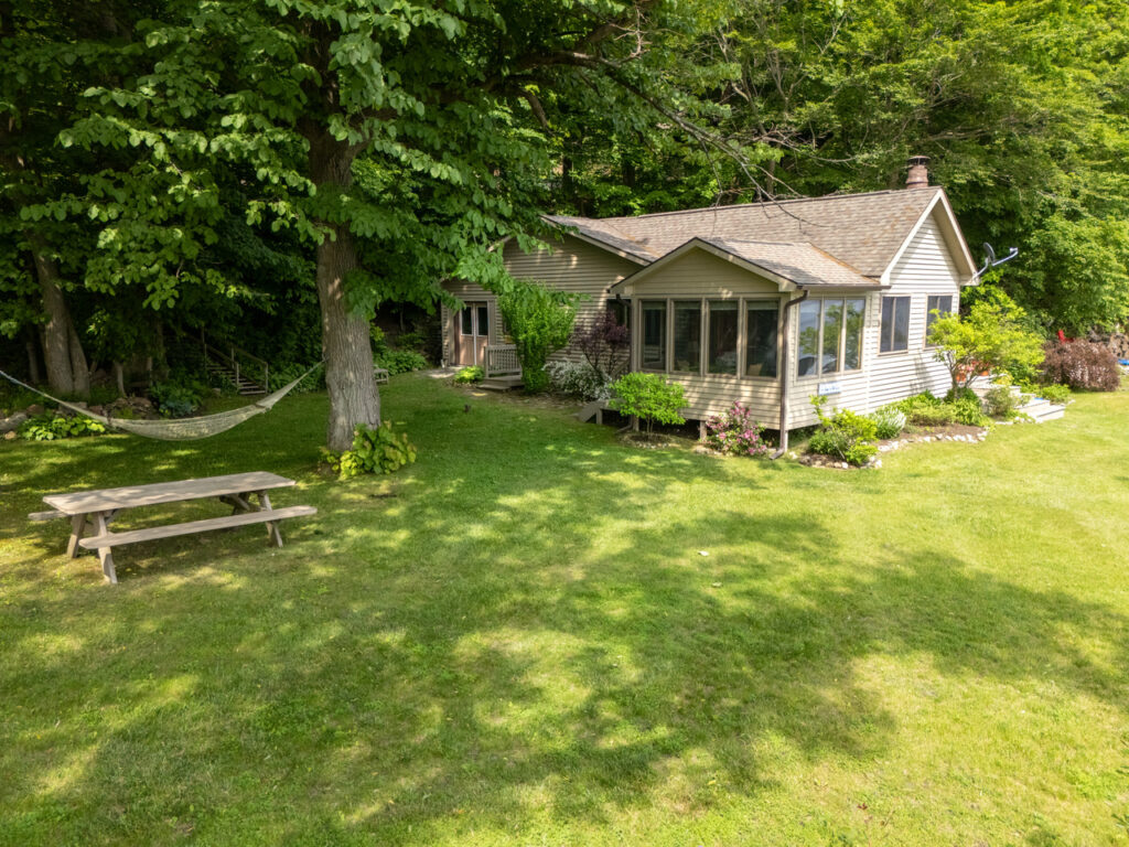 Another view of the cottage, which is on the viewer's right, and a picnic table and hammock on the viewer's left. A tree stands between the cottage and the hammock. It is a suspension point for the hammock.