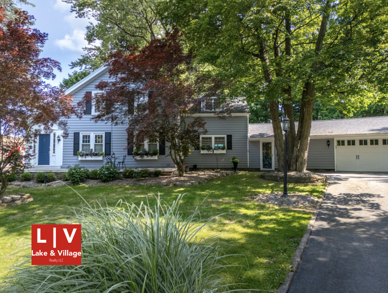 This image shows a light blue house with blue shutters at the edge of a lawn and driveway. A large tree with green leaves and a smaller tree with dark red leaves stand in front of the house. The red-and-white Lake and Village logo is in the viewer's lower left-hand corner.