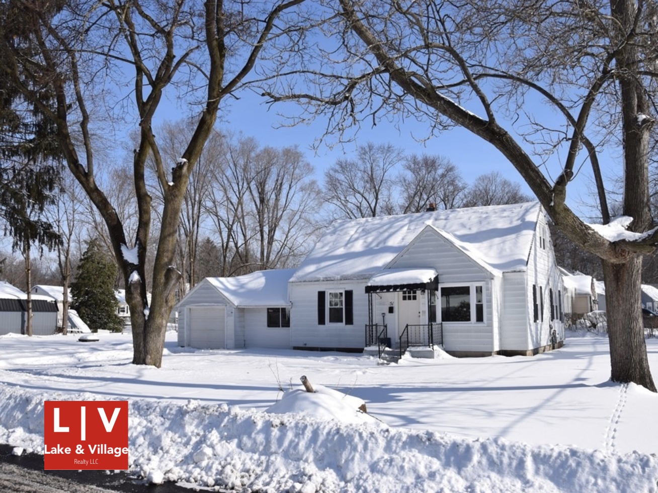 This image shows a white-sided home in snow. Two trees, mostly bare, stand in the front yard. More bare trees stand behind the house. The sky is blue. The red-and-white Lake and Village logo is in the viewer’s lower left-hand corner.